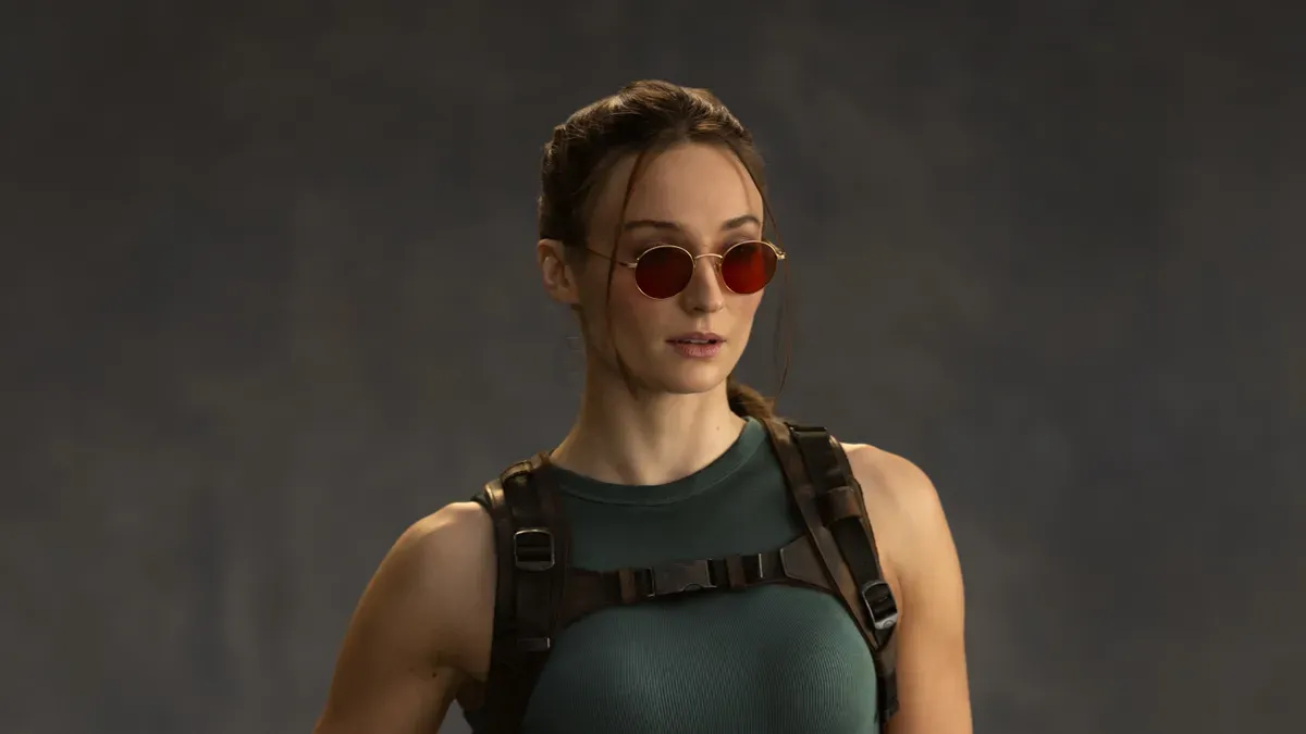 Sophie Turner as Lara Croft in Prime Video's Tomb Raider series, wearing a game-accurate costume with pistols and shorts, set against a rugged outdoor backdrop.